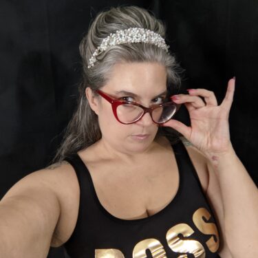 Grey haired older woman holds her red eyeglass frames and looks over her glasses at you. She is wearing a tiara and a tank top that says BOSS in gold.
