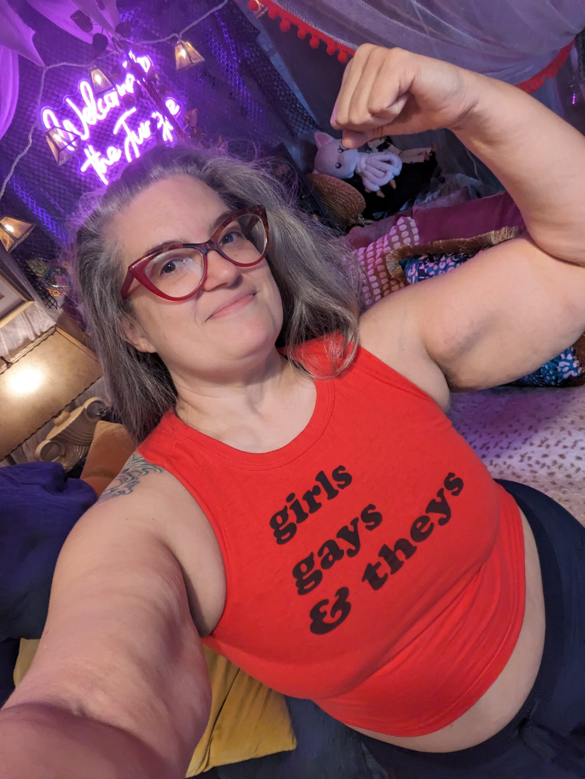 OctoGoddess flexes her left bicep and wears a tight red tank top that reads "girls, gays and theys"
