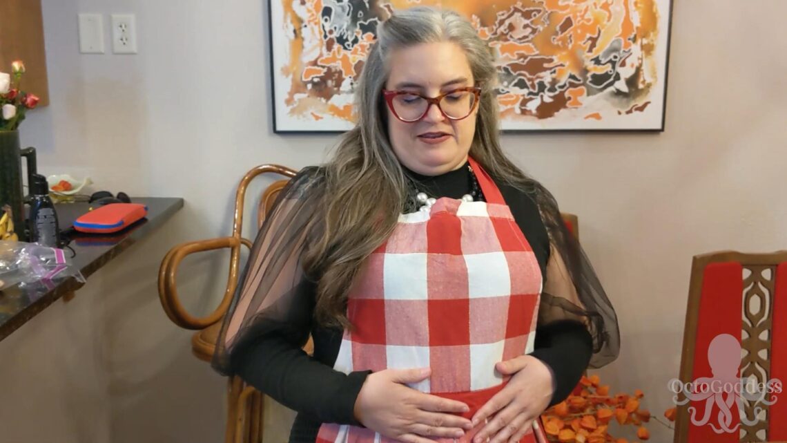 Woman with long hair, looks down while rubbing her belly. She wears a red checked apron.